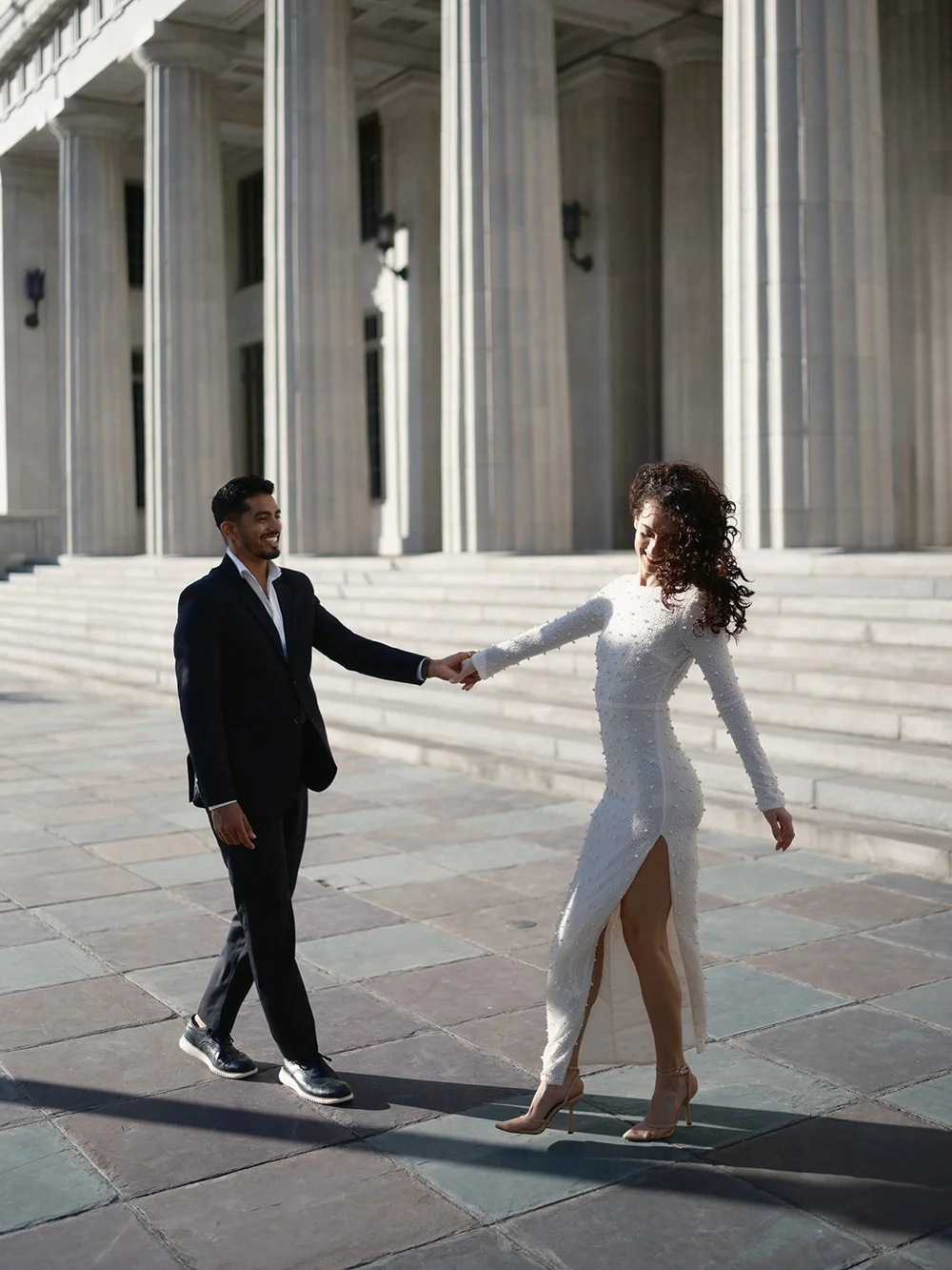 Groom holding the bride's hand as they walk across the courthouse plaza in strong afternoon light
