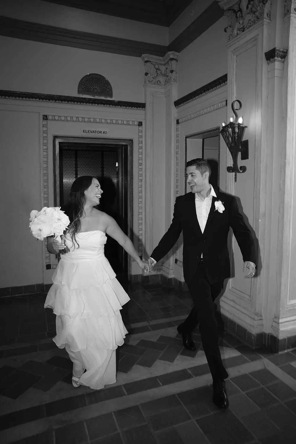 Black and white photo of Chris and David walking down a hallway, she carrying her white bouquet