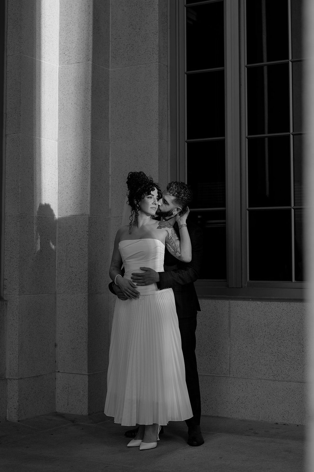 Black and white photo of the bride and groom embracing beside tall courthouse windows