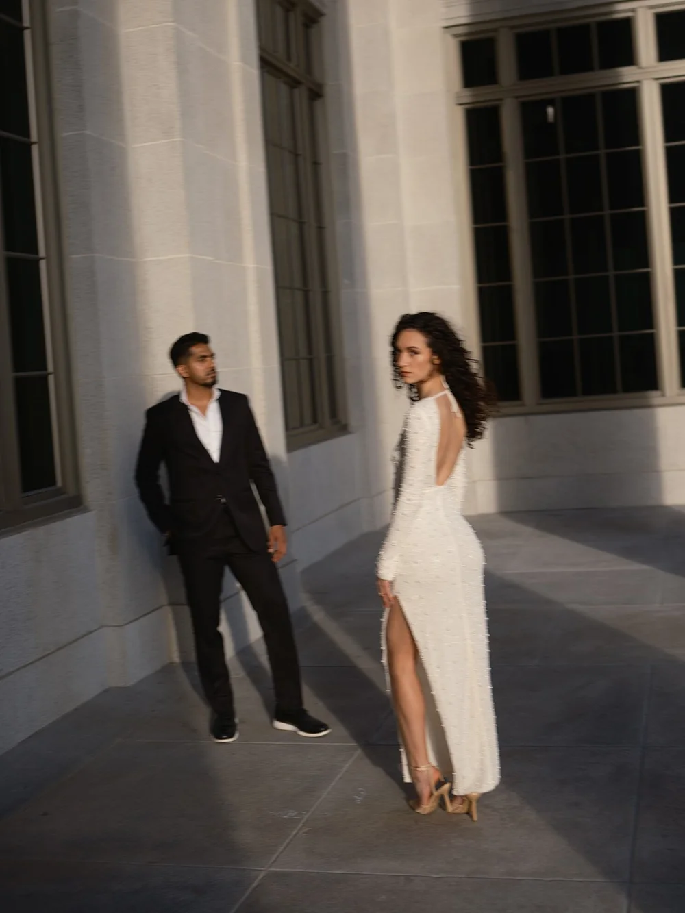 Groom leaning on a stone wall and the bride in a white dress standing apart beside tall courthouse windows