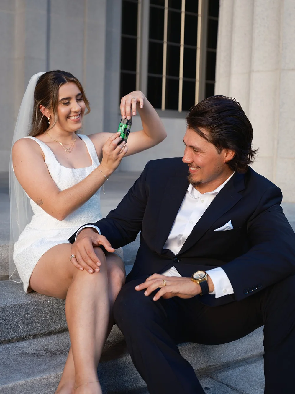 Bride laughing and looking at a small bottle in her hand while the groom sits beside her on the steps