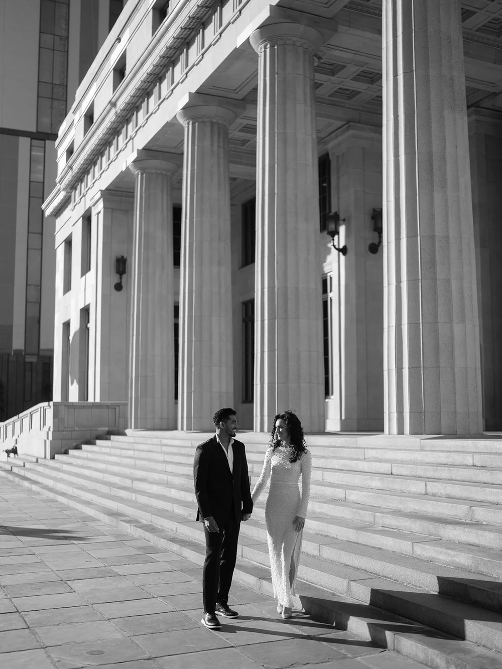 Bride and groom walking together across the wide stone steps in front of the courthouse columns