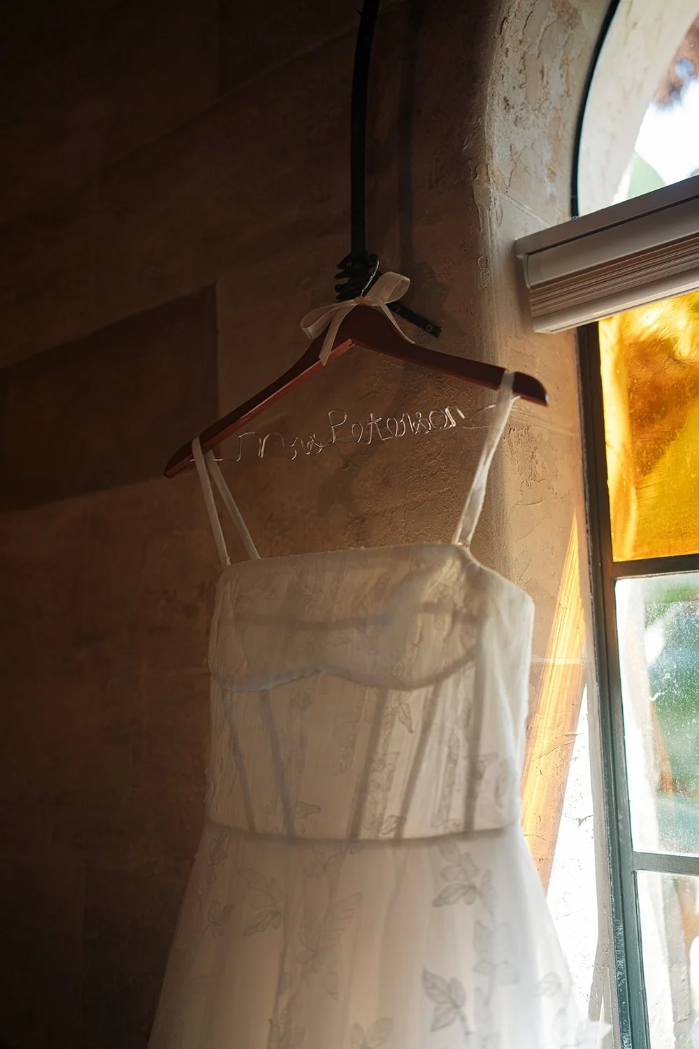 Wedding dress hanging on a wooden hanger beside a window with bright light coming in