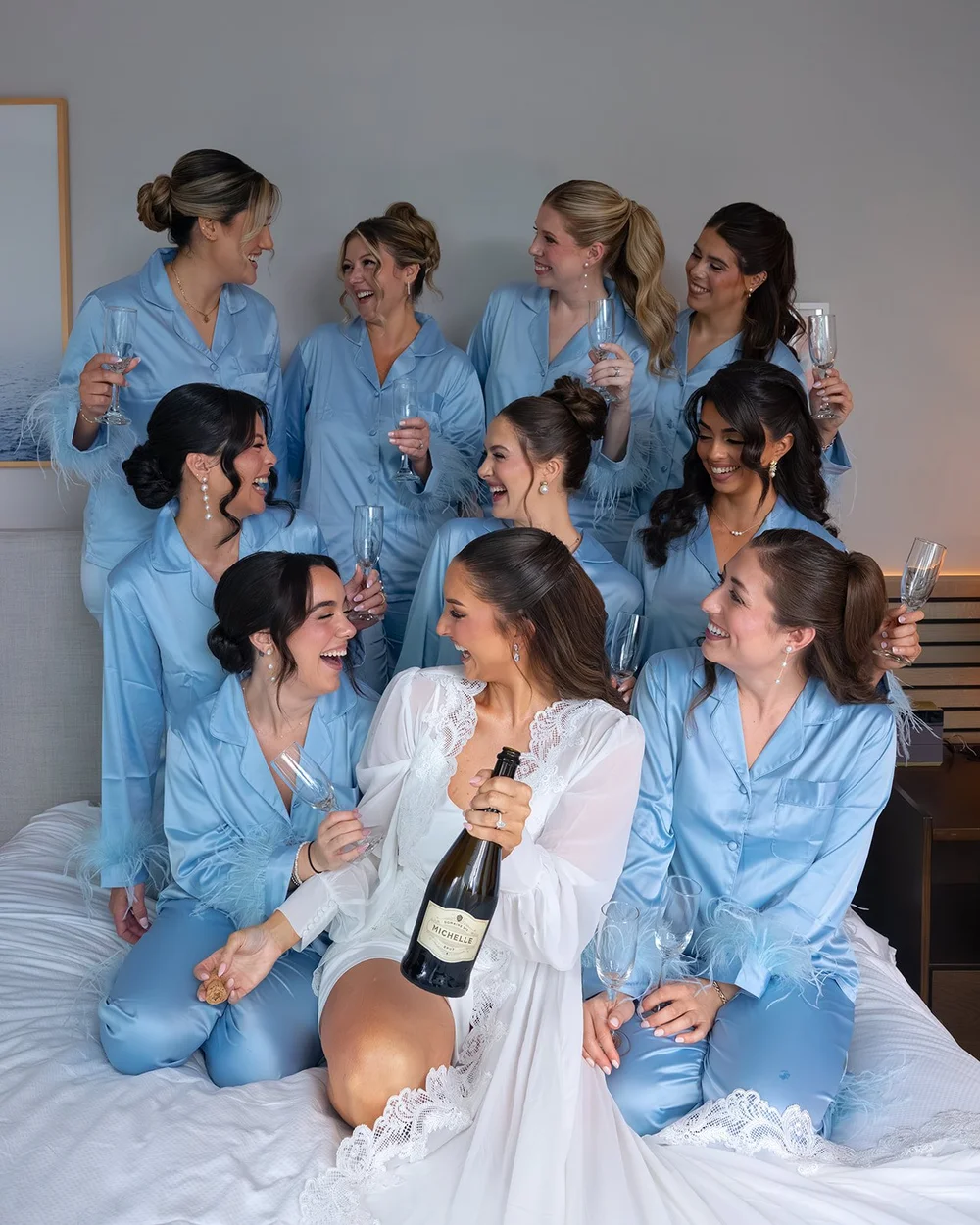Rachel in a white robe surrounded by bridesmaids in matching light blue robes, laughing on the bed with a bottle of champagne