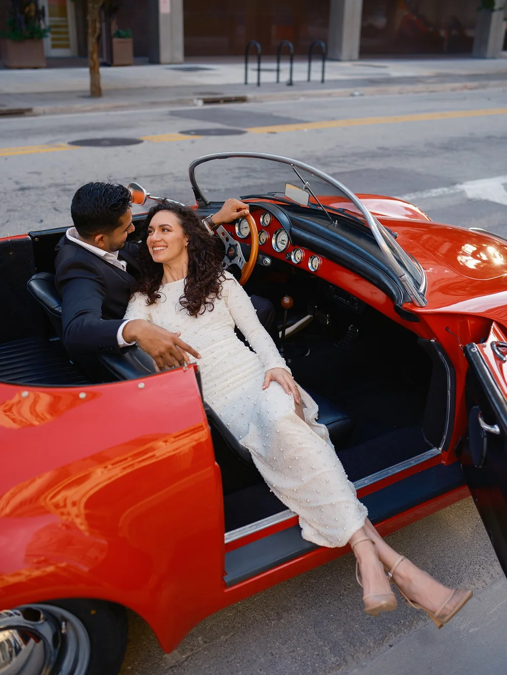 Bride sitting sideways in the open door of the red convertible with the groom beside her