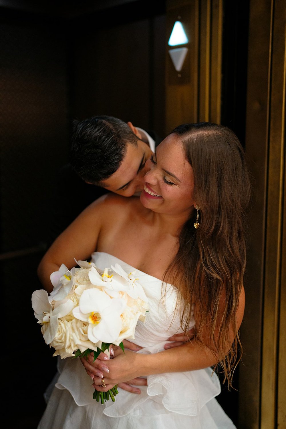 Chris laughing with her white bouquet while David kisses her cheek in a dark stairwell