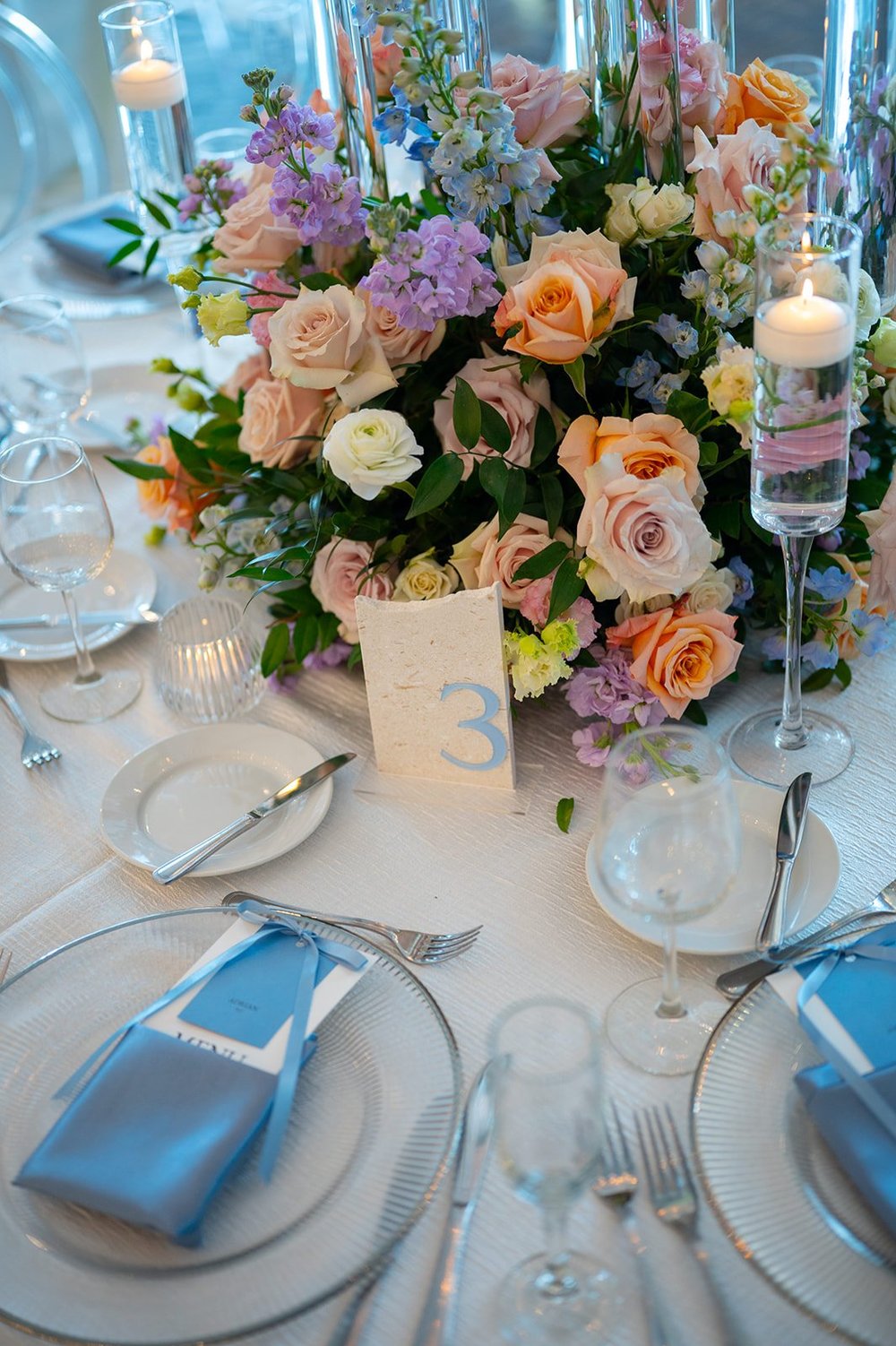 Reception table with a pastel flower centerpiece, candles and a place setting with a blue napkin