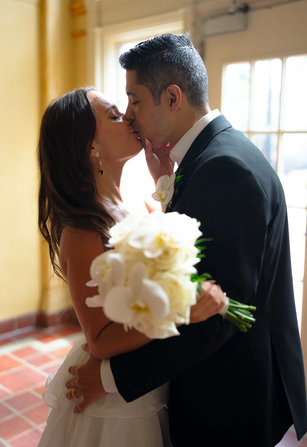 Close kiss between Chris and David in warm window light with the white roses of her bouquet in front