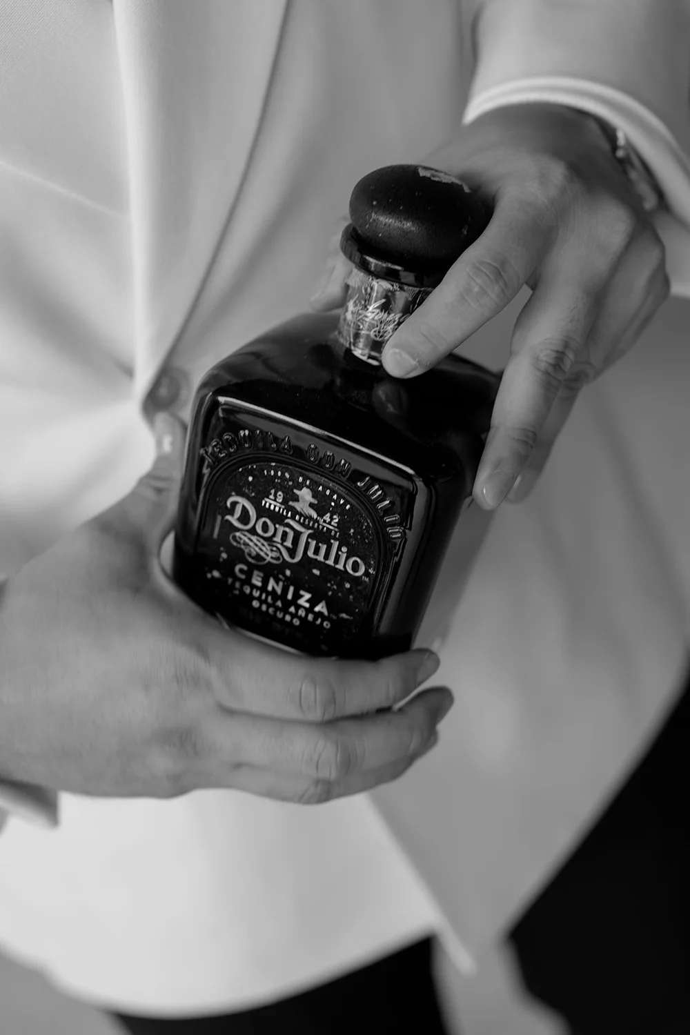 Black and white detail of a hand holding a bottle of Don Julio tequila in front of a white jacket