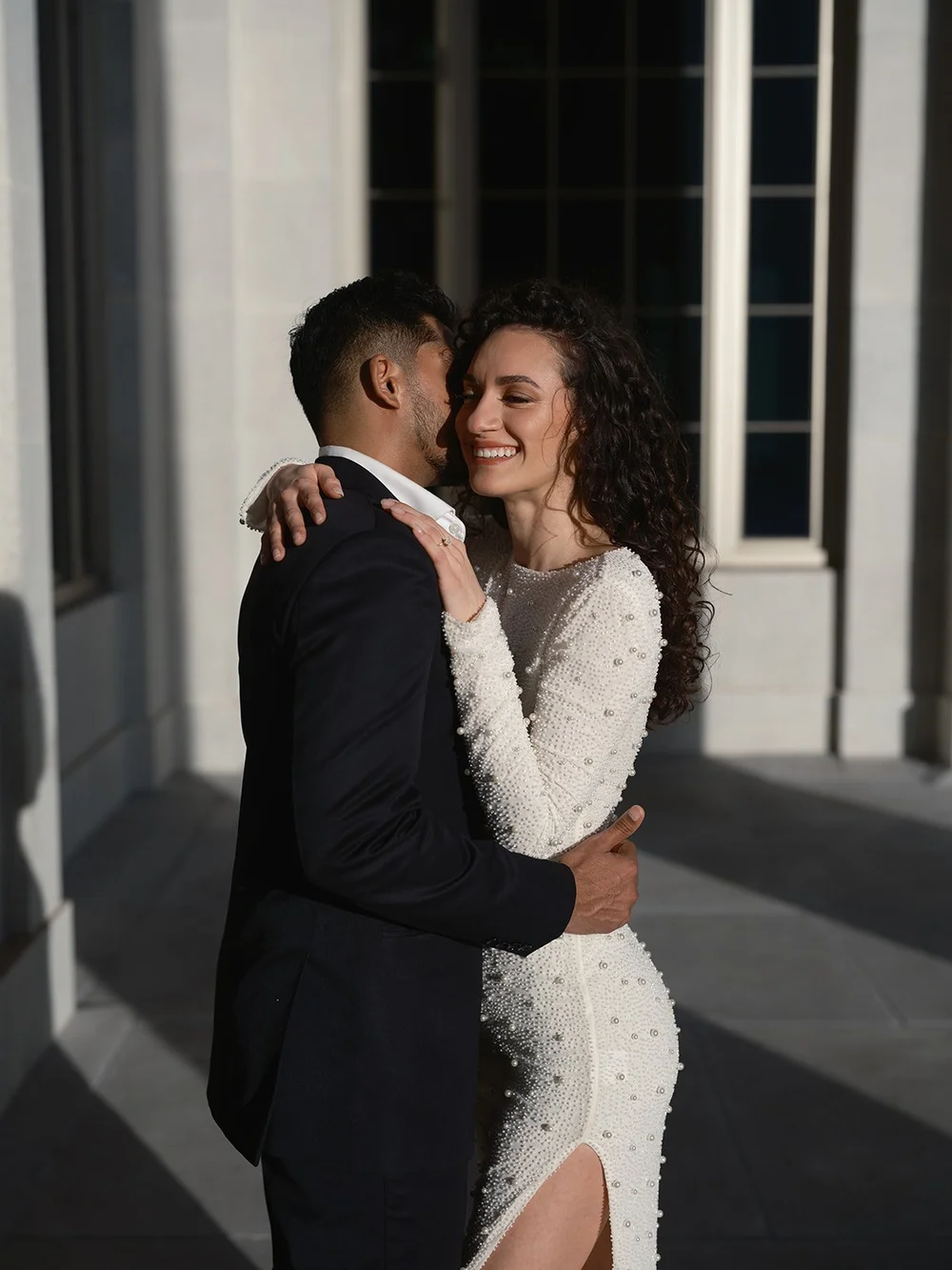 Bride and groom embracing in the sun next to a tall window of the courthouse