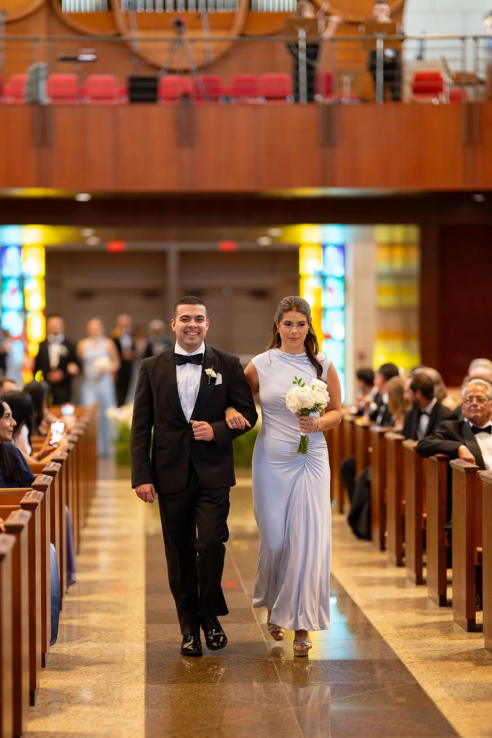 A bridesmaid in a light blue dress walking down the church aisle with a groomsman in a black tuxedo