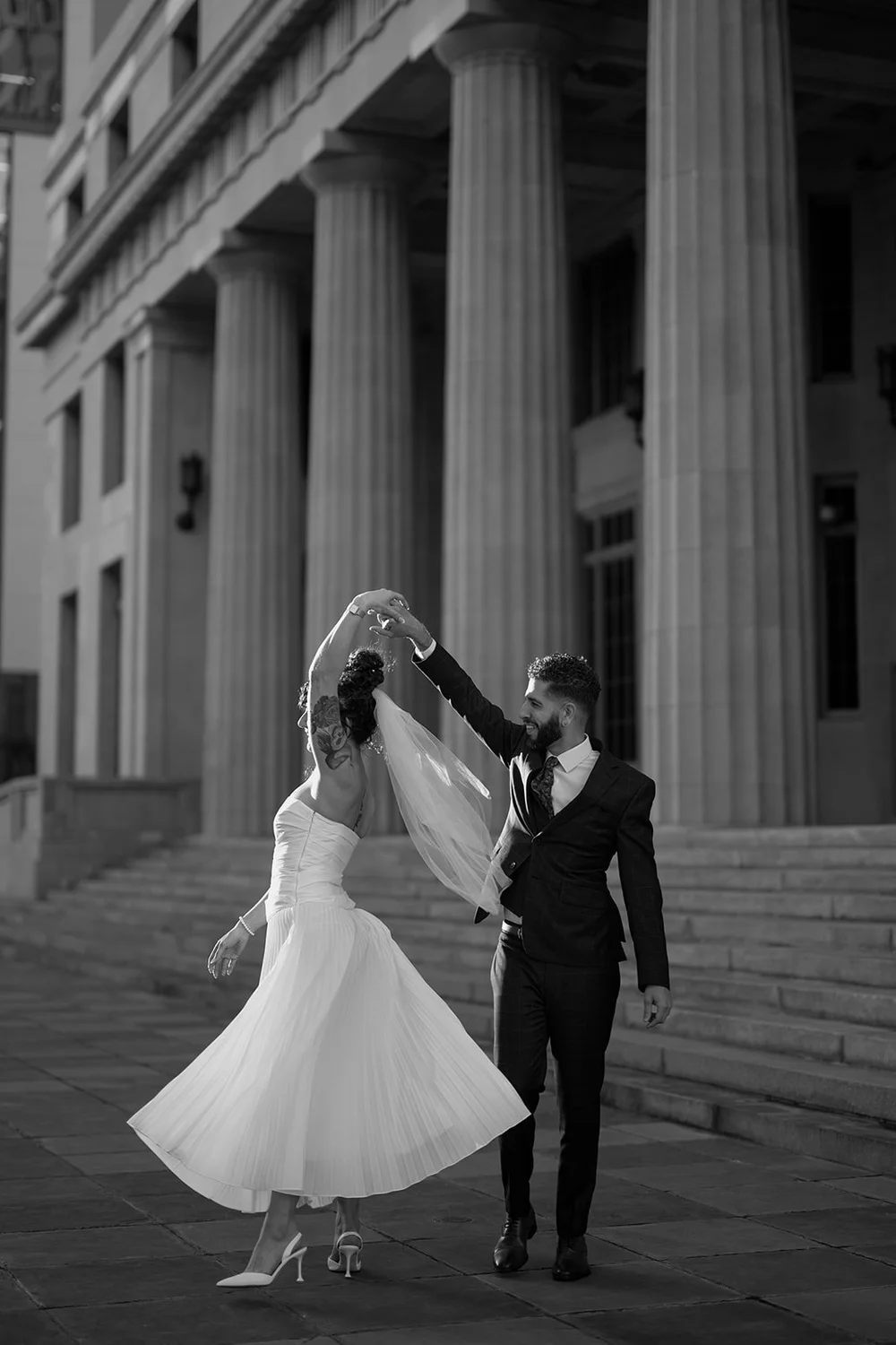 Black and white photo of the bride twirling in her white dress while the groom holds her hand in front of tall courthouse columns