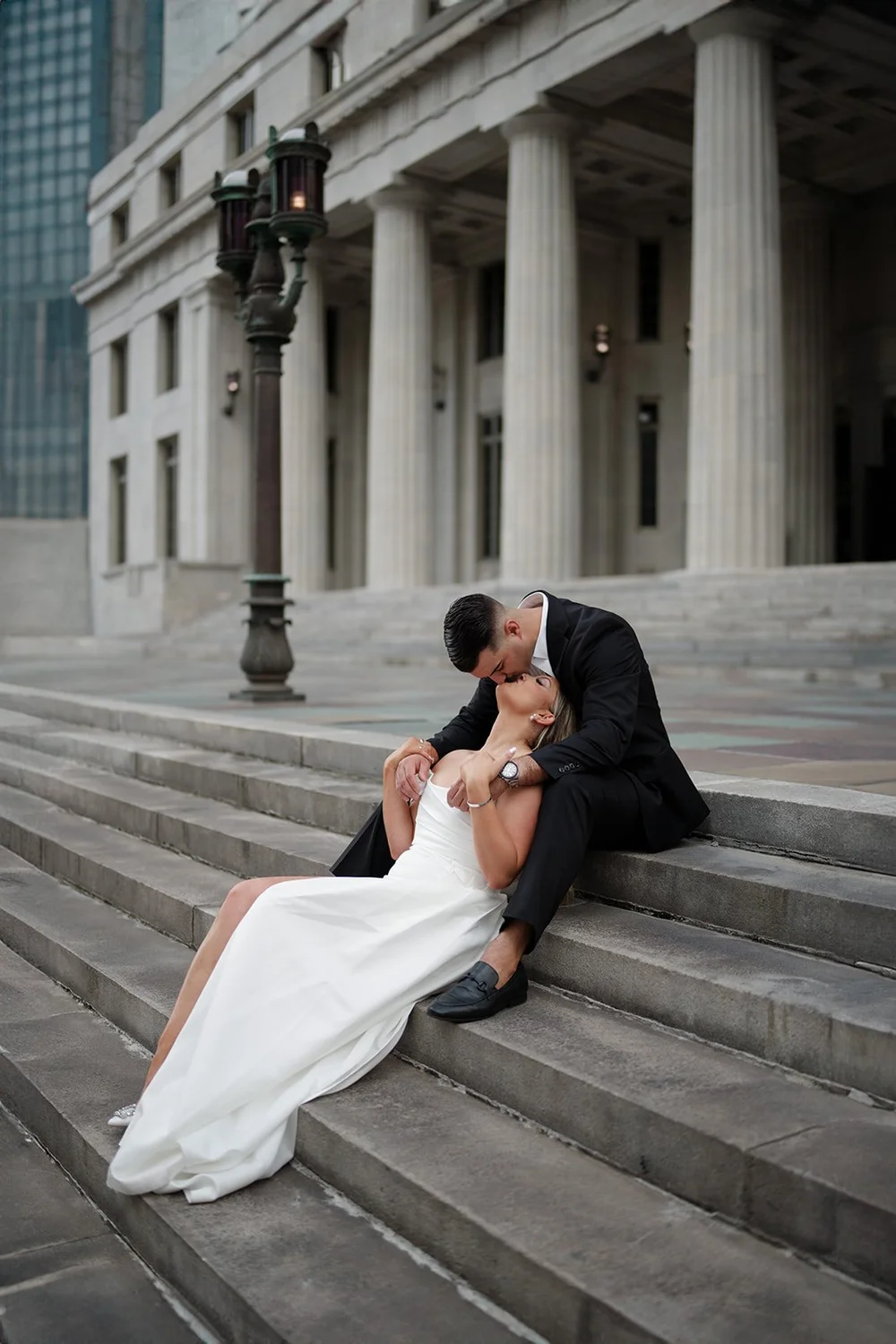 Rachel and Austin sitting on the courthouse steps in a kiss with a street lamp and downtown towers behind them