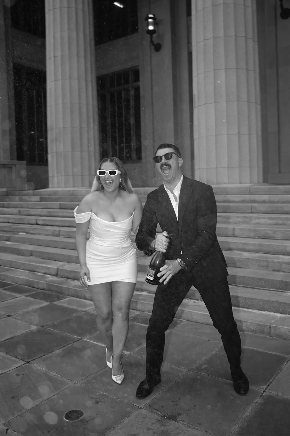 Austin holding a champagne bottle while Rachel laughs beside him on the courthouse steps, in black and white