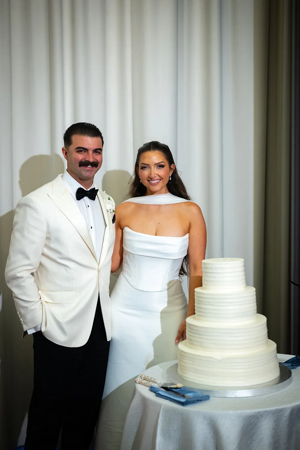 Rachel and Austin smiling together beside their tiered white wedding cake