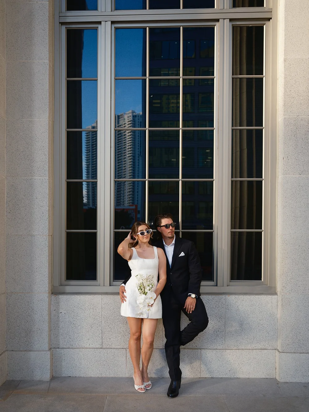 Bride and groom standing in front of the courthouse's tall windows with blue sky reflected in the glass