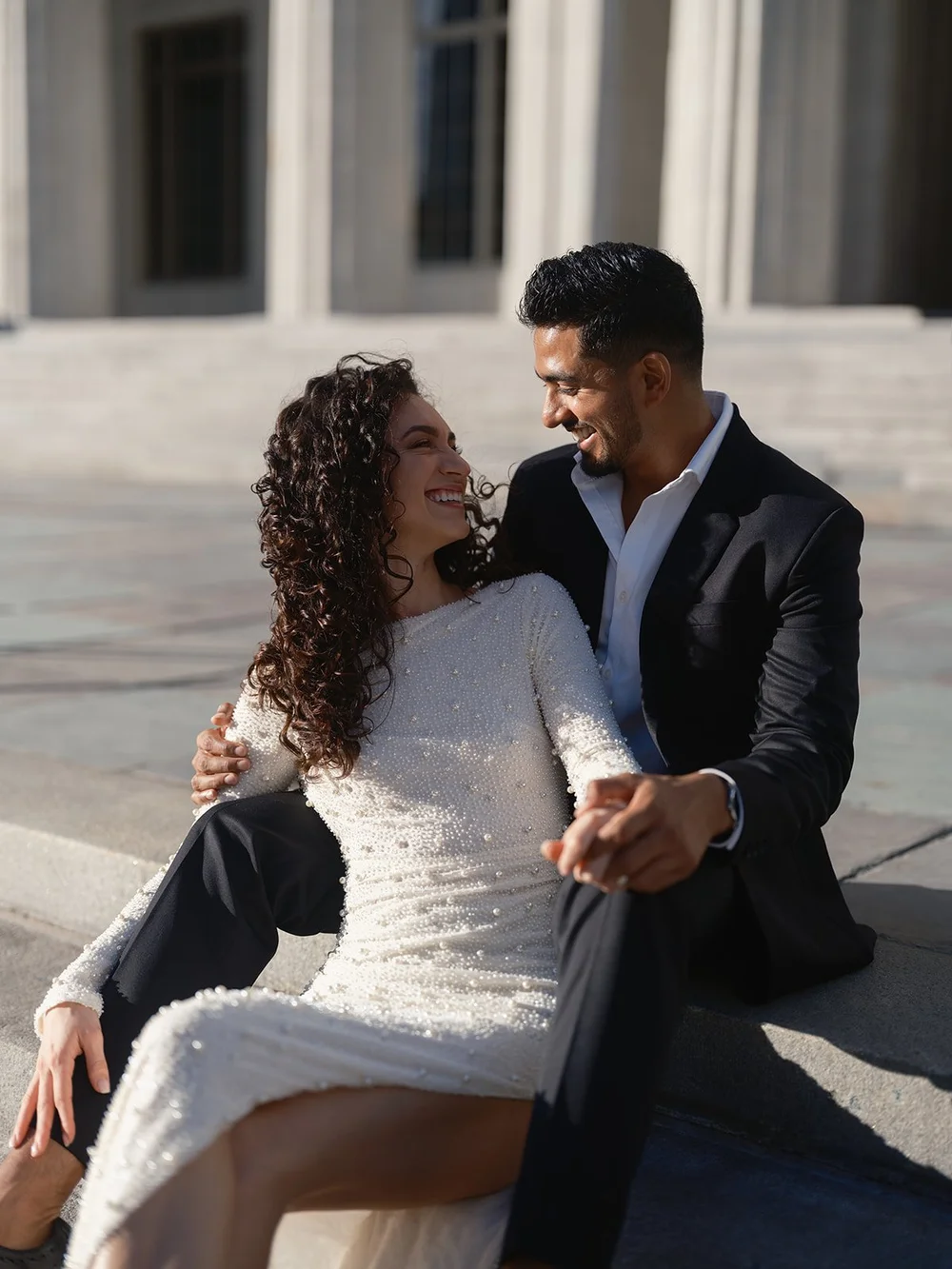 Black and white photo of the bride and groom sitting close together on sunlit steps, smiling at each other