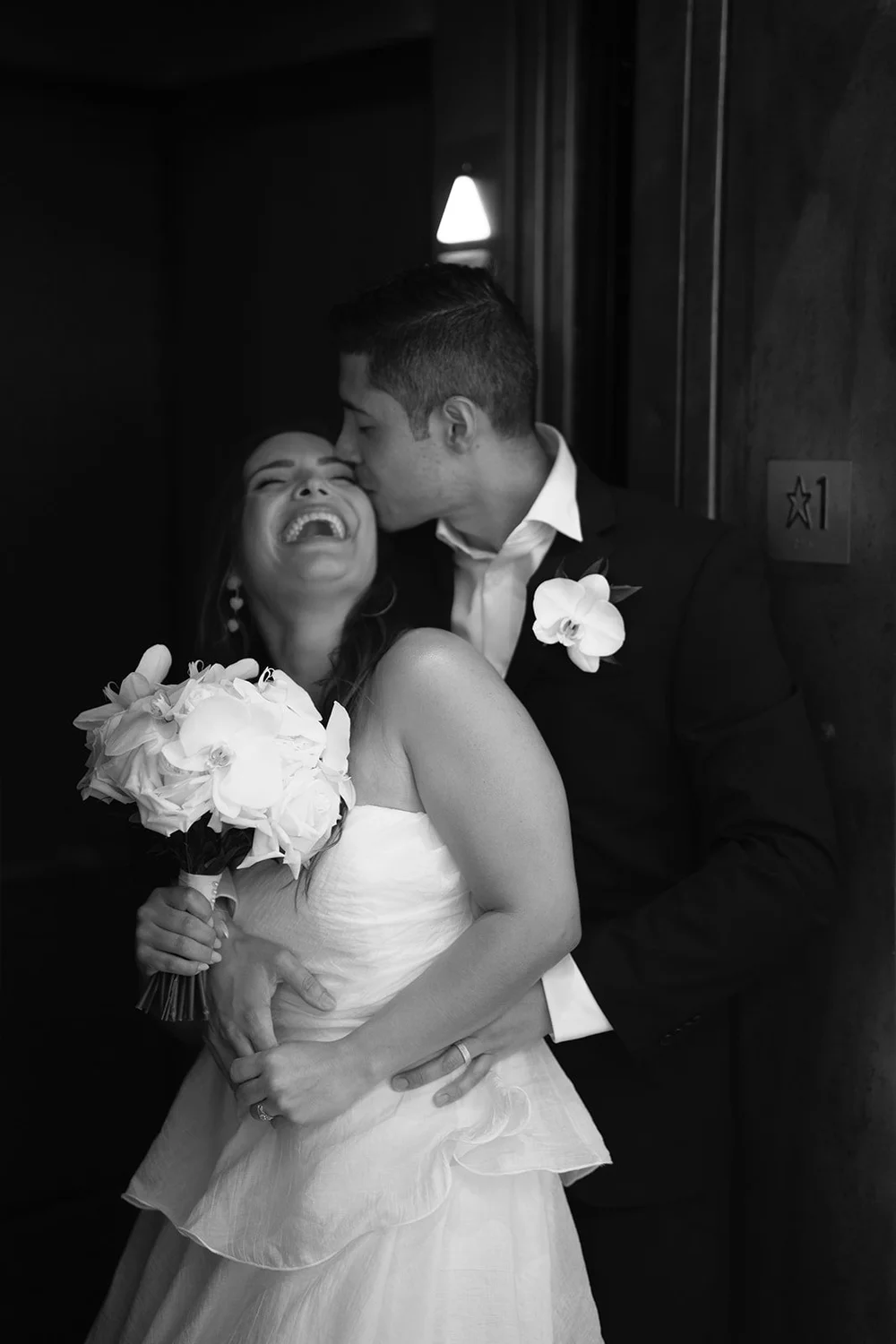Black and white flash portrait of Chris laughing while David kisses her cheek, her bouquet at her waist
