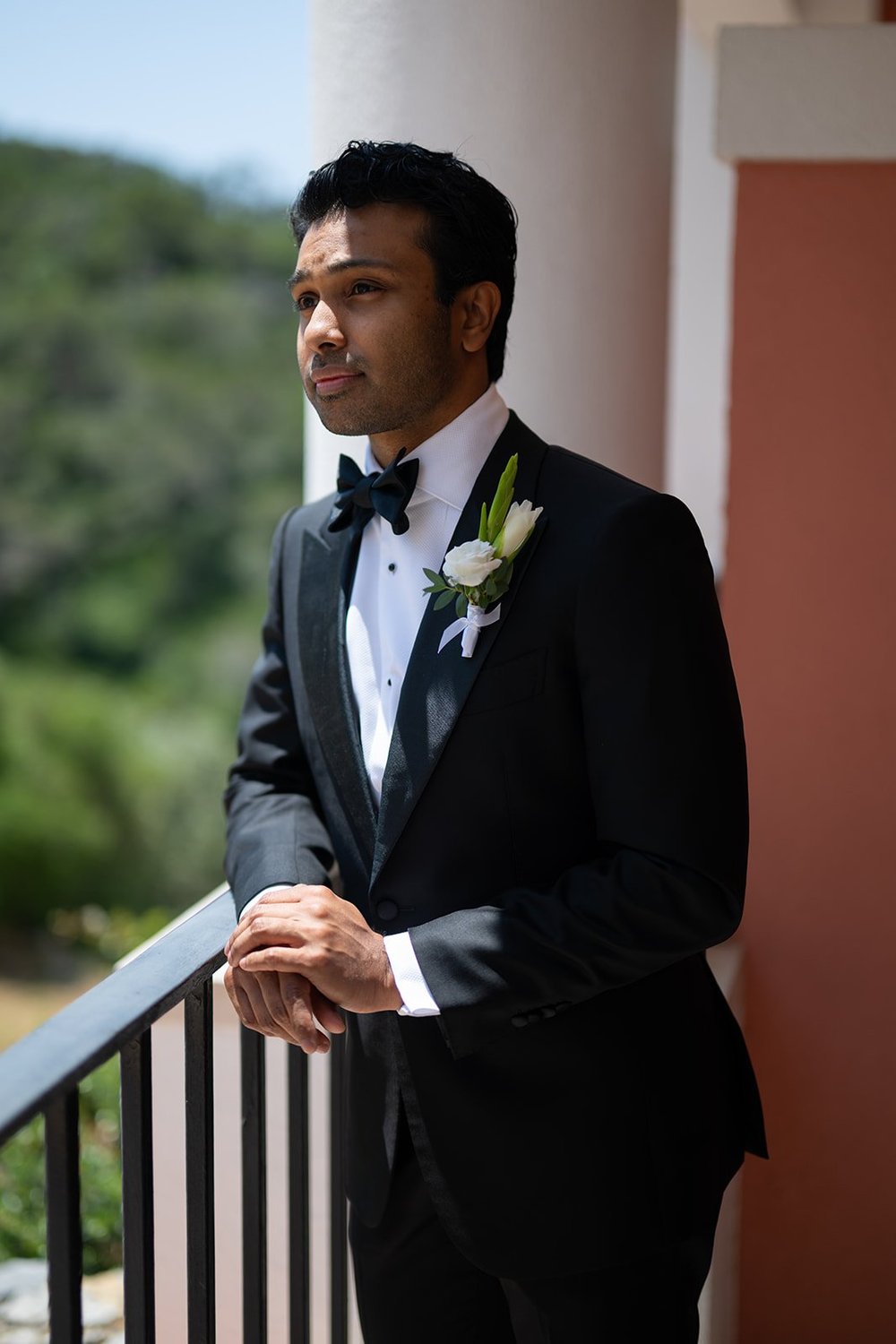 Jacob in a tuxedo with a white boutonniere leaning on a balcony railing with green hills behind him