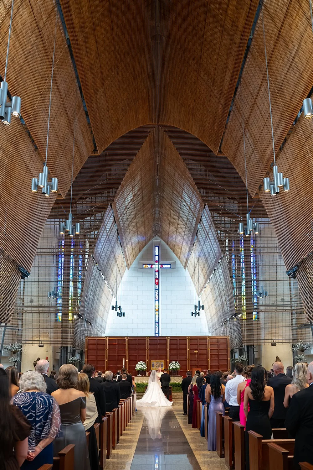 Rachel and Austin at the altar at the end of a long aisle under the wood ceiling of Church of the Epiphany in Miami, with guests filling the pews