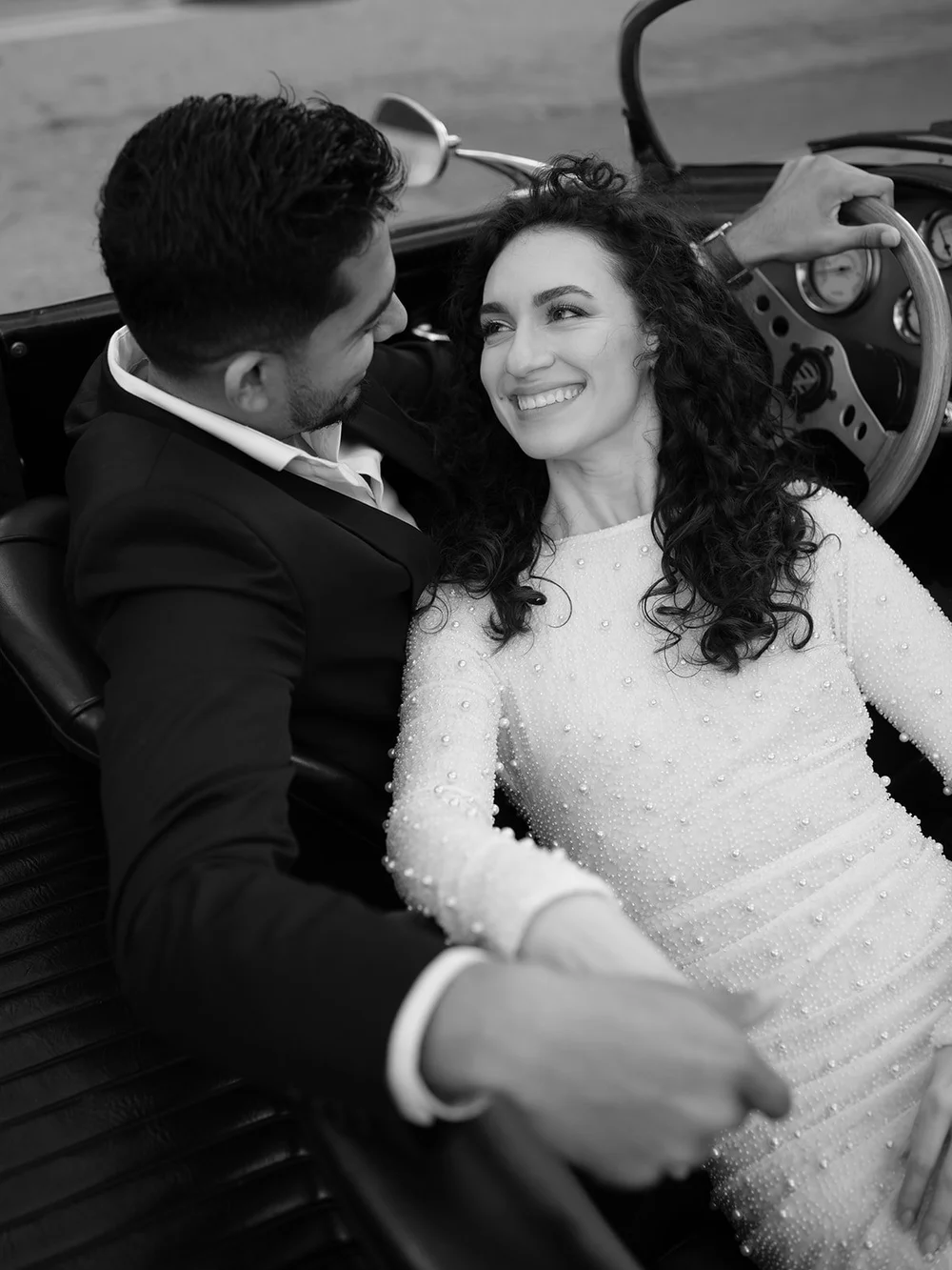 Black and white photo of the bride smiling at the camera as the groom leans toward her in the red convertible