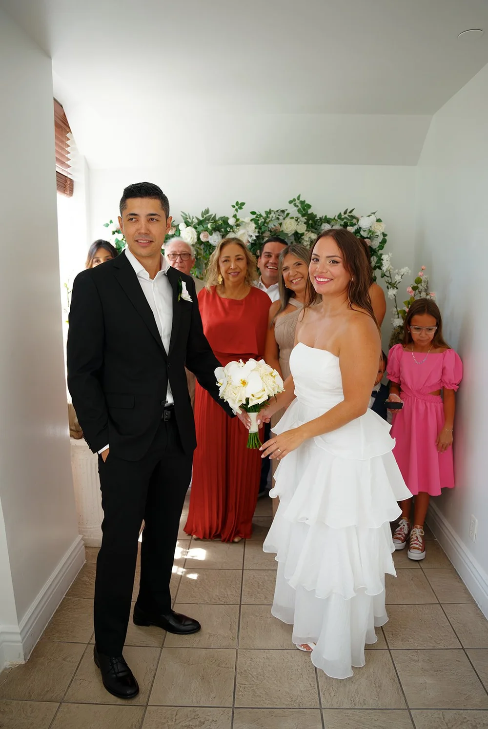 Chris and David standing side by side with family and a girl in pink along a white wall with flowers
