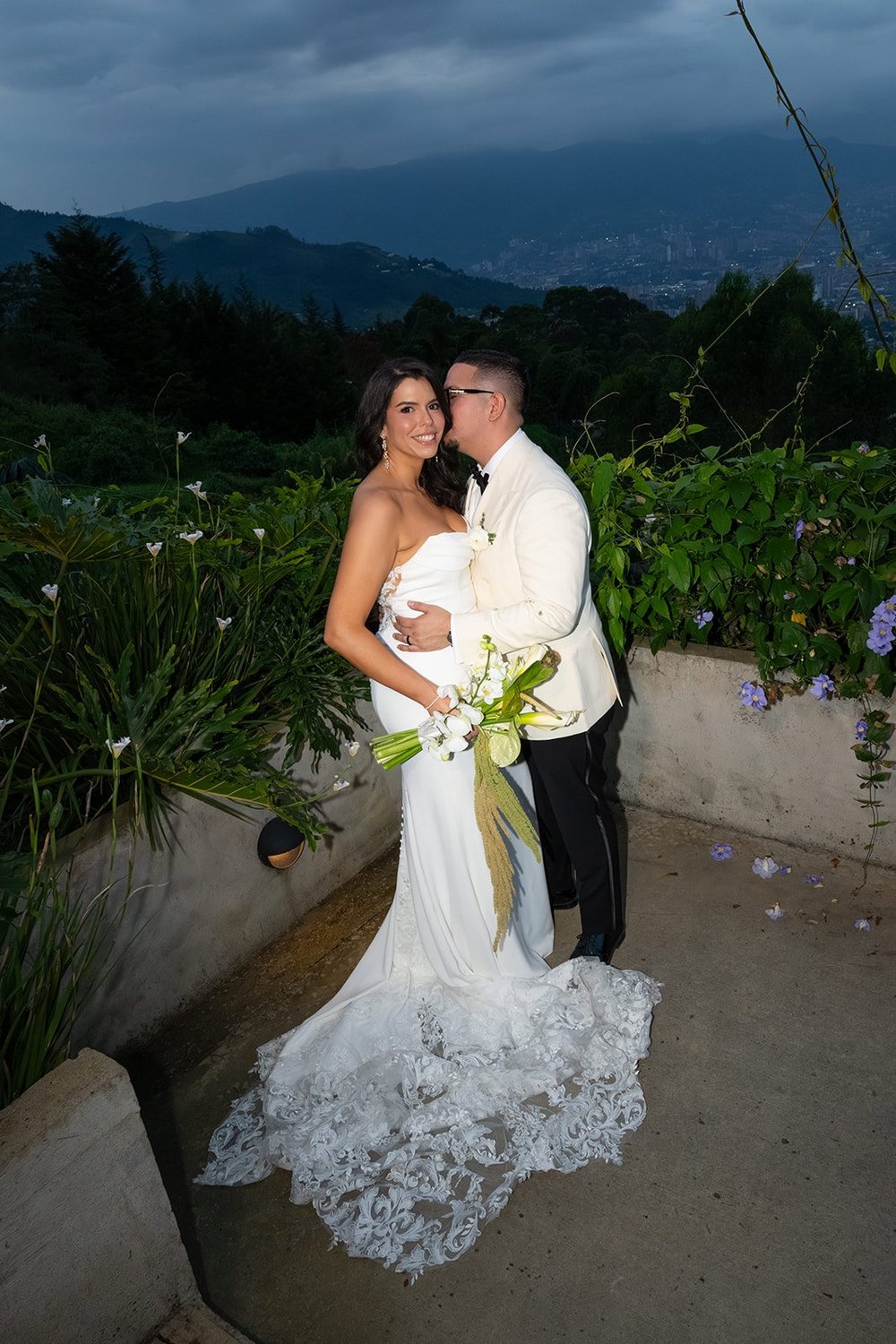 Krystine smiling at the camera as Douglas kisses her cheek on a terrace at dusk, with the Medellín valley and cloudy mountains behind them