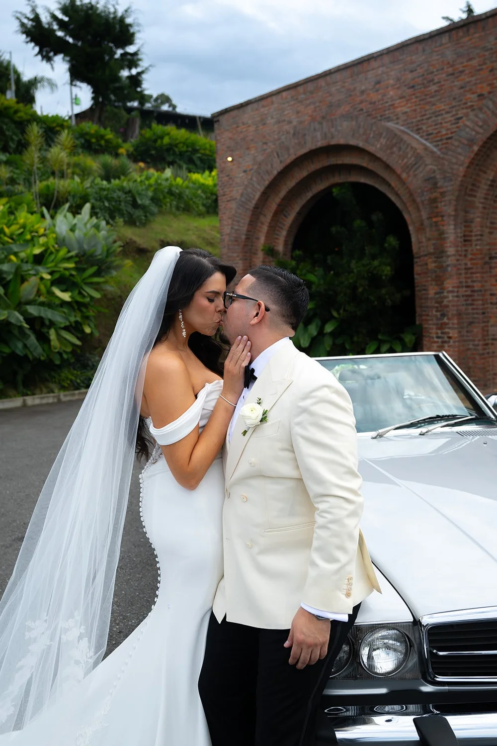 Krystine and Douglas kissing beside a white vintage Mercedes in front of a brick arch at Voilà Eventos in Medellín, Colombia