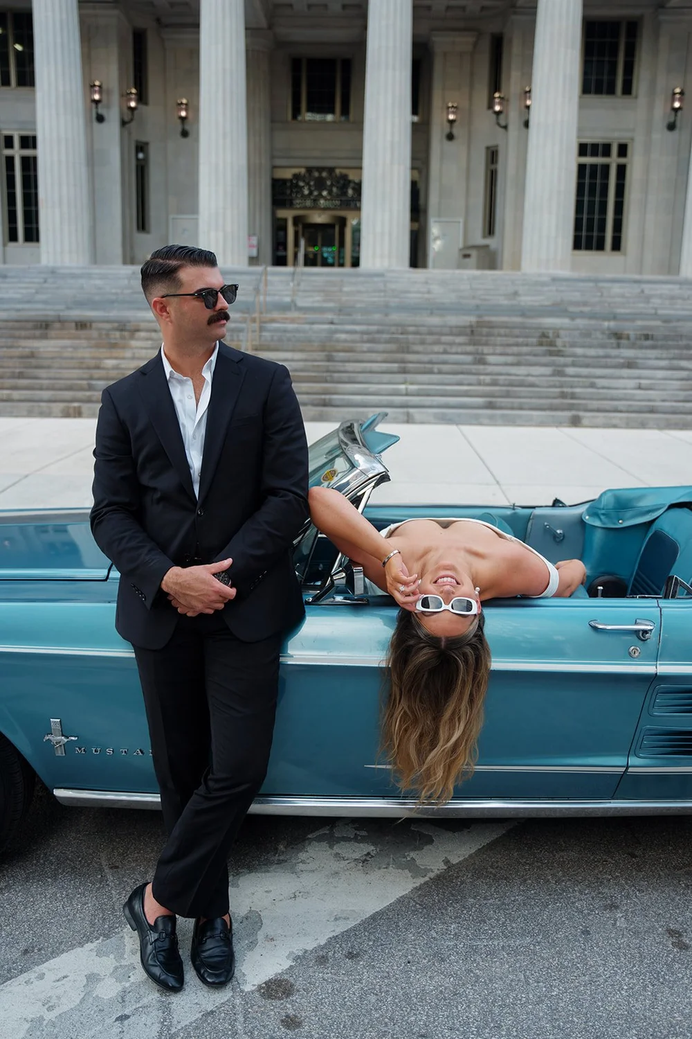 Austin standing in a black suit and sunglasses while Rachel lies back across the Mustang's trunk