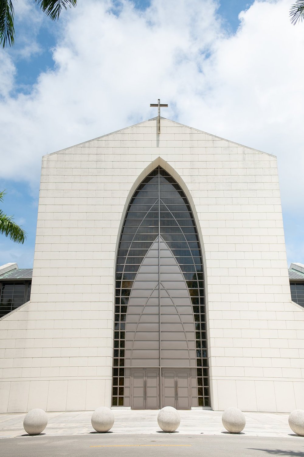 Front of the church with a tall pointed arch window and a cross against a blue sky