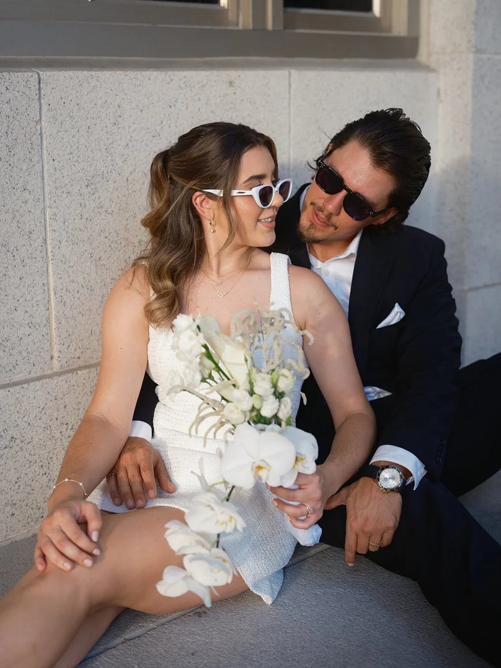 Bride in white sunglasses holding a white orchid bouquet while the groom in sunglasses leans in beside her