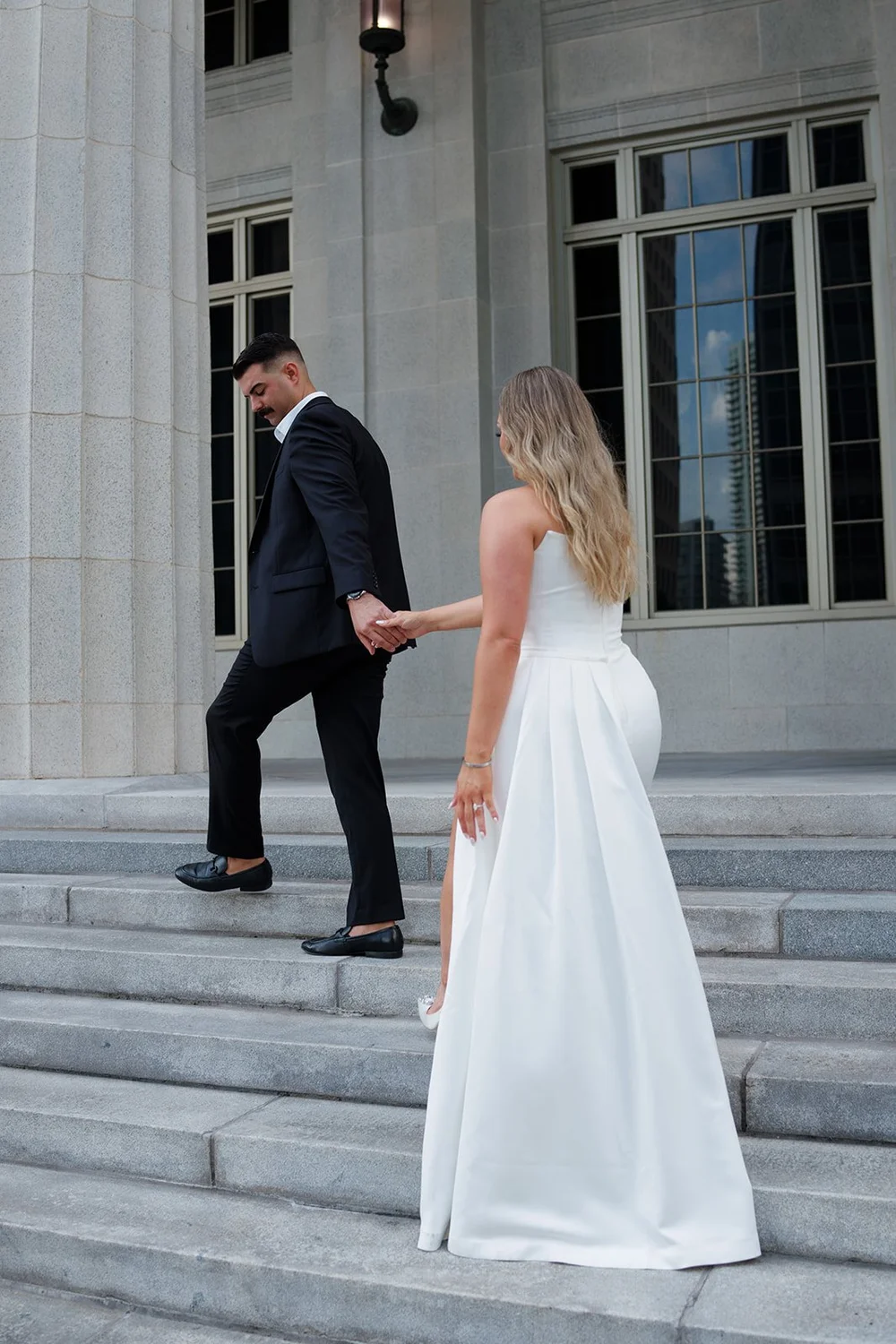 Austin and Rachel holding hands as they walk up the courthouse steps, Rachel in a long white gown