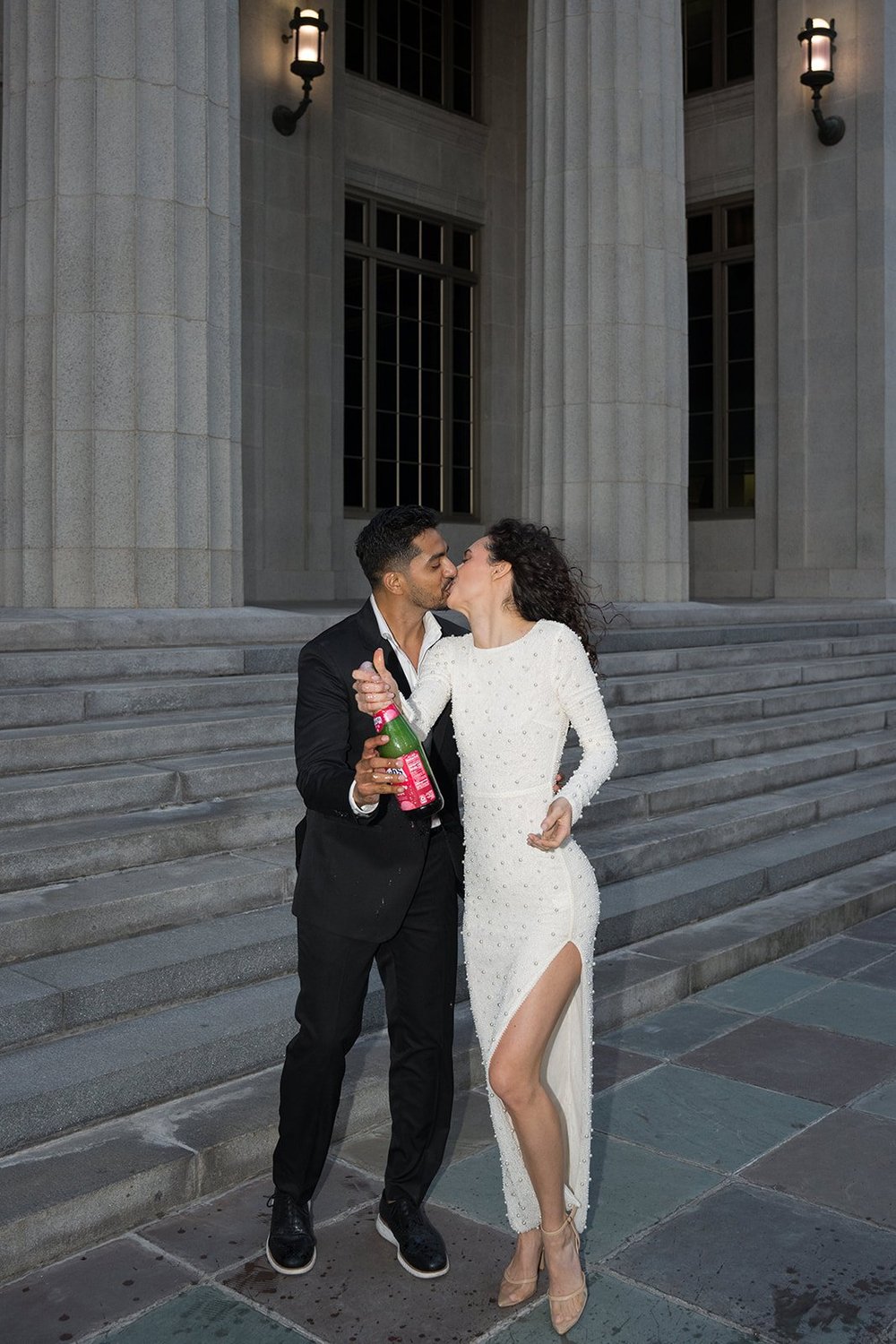 Groom and bride kissing on the courthouse steps at dusk, with a bottle in his hand and lamps lit behind them