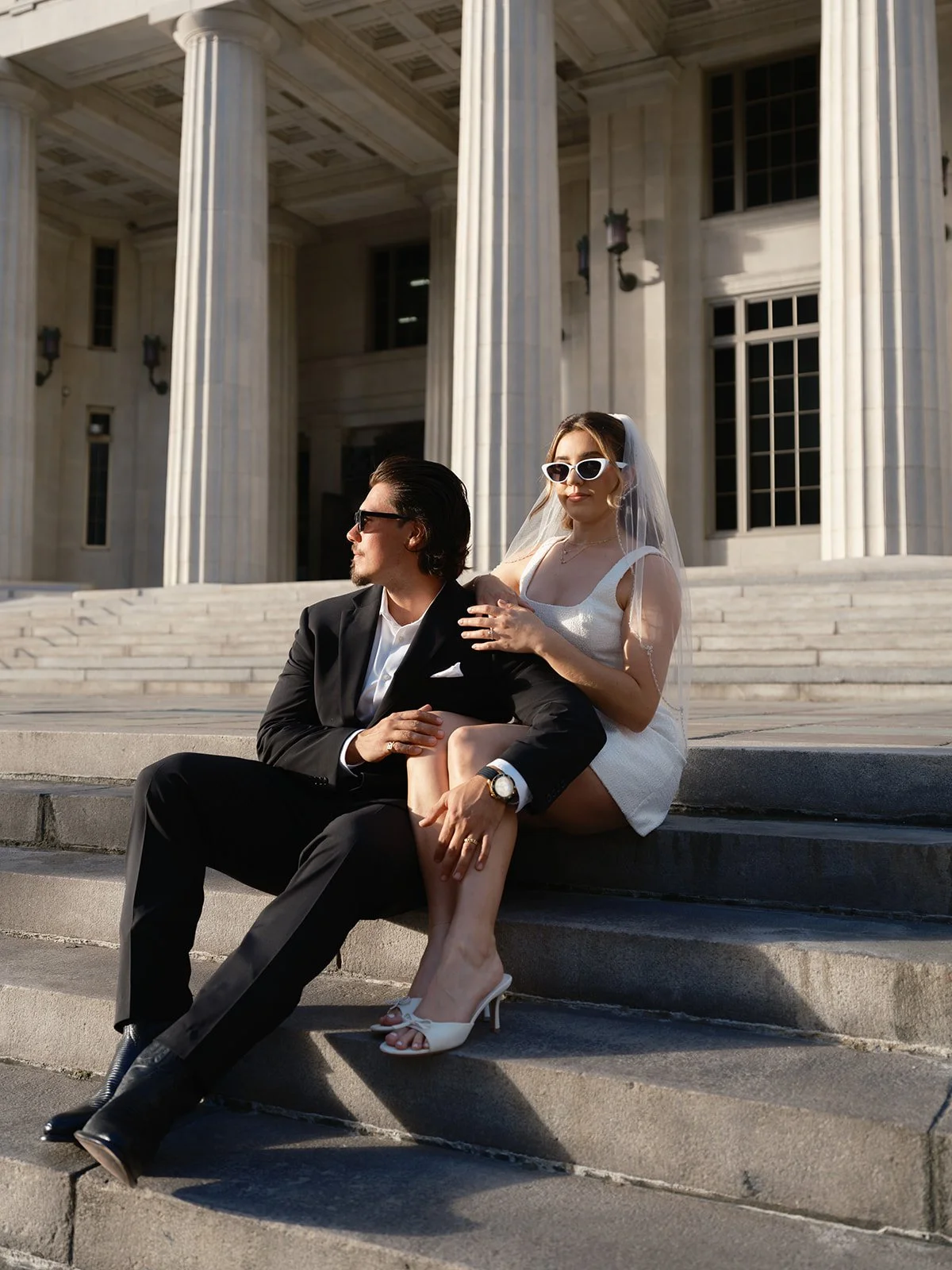 Carina and Billy sitting on the steps of the Miami-Dade County Courthouse under the tall columns, she in sunglasses, a short white dress and a veil, for their Miami courthouse elopement