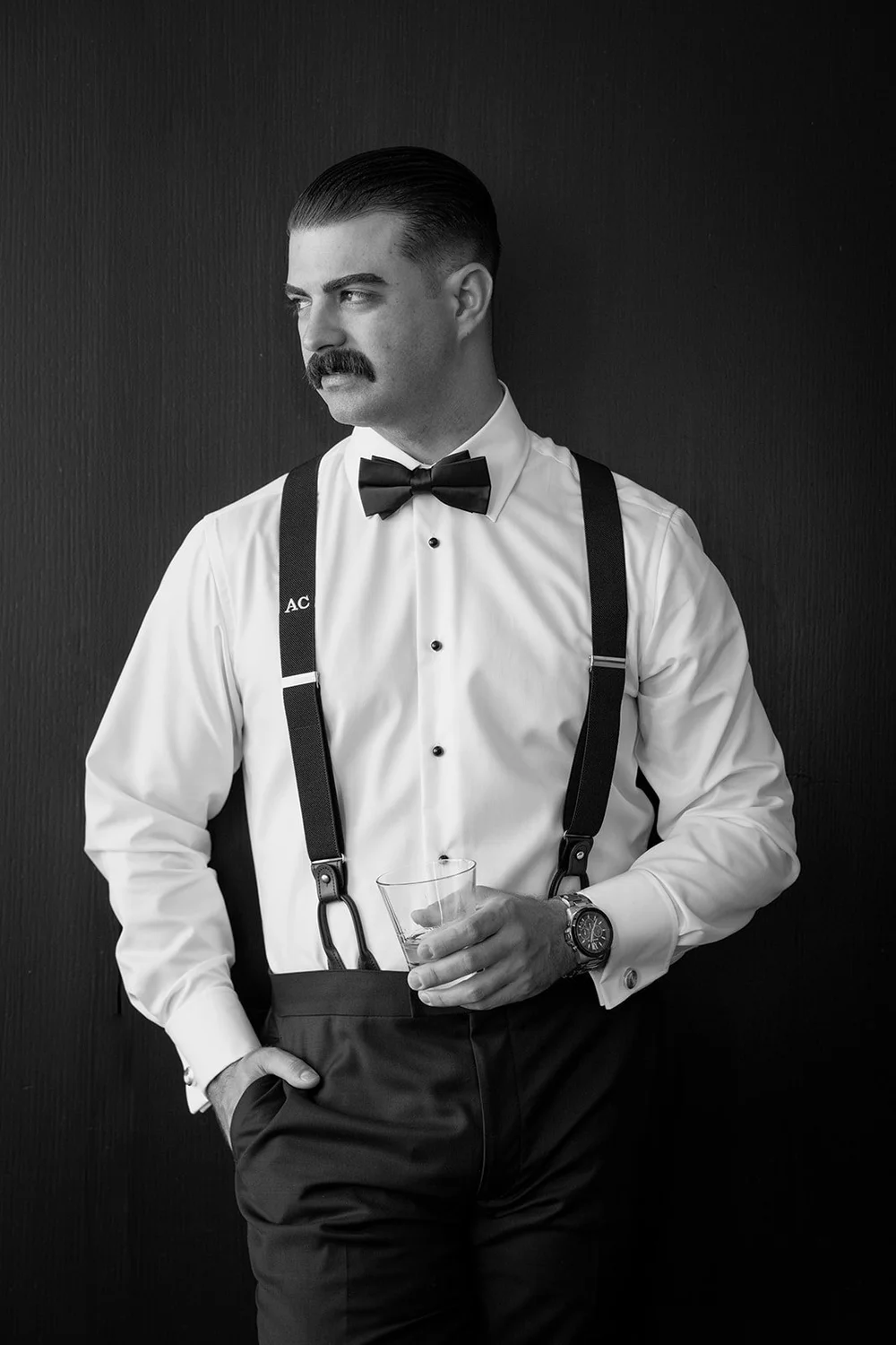 Austin in suspenders and a bow tie standing against a dark wall, in black and white