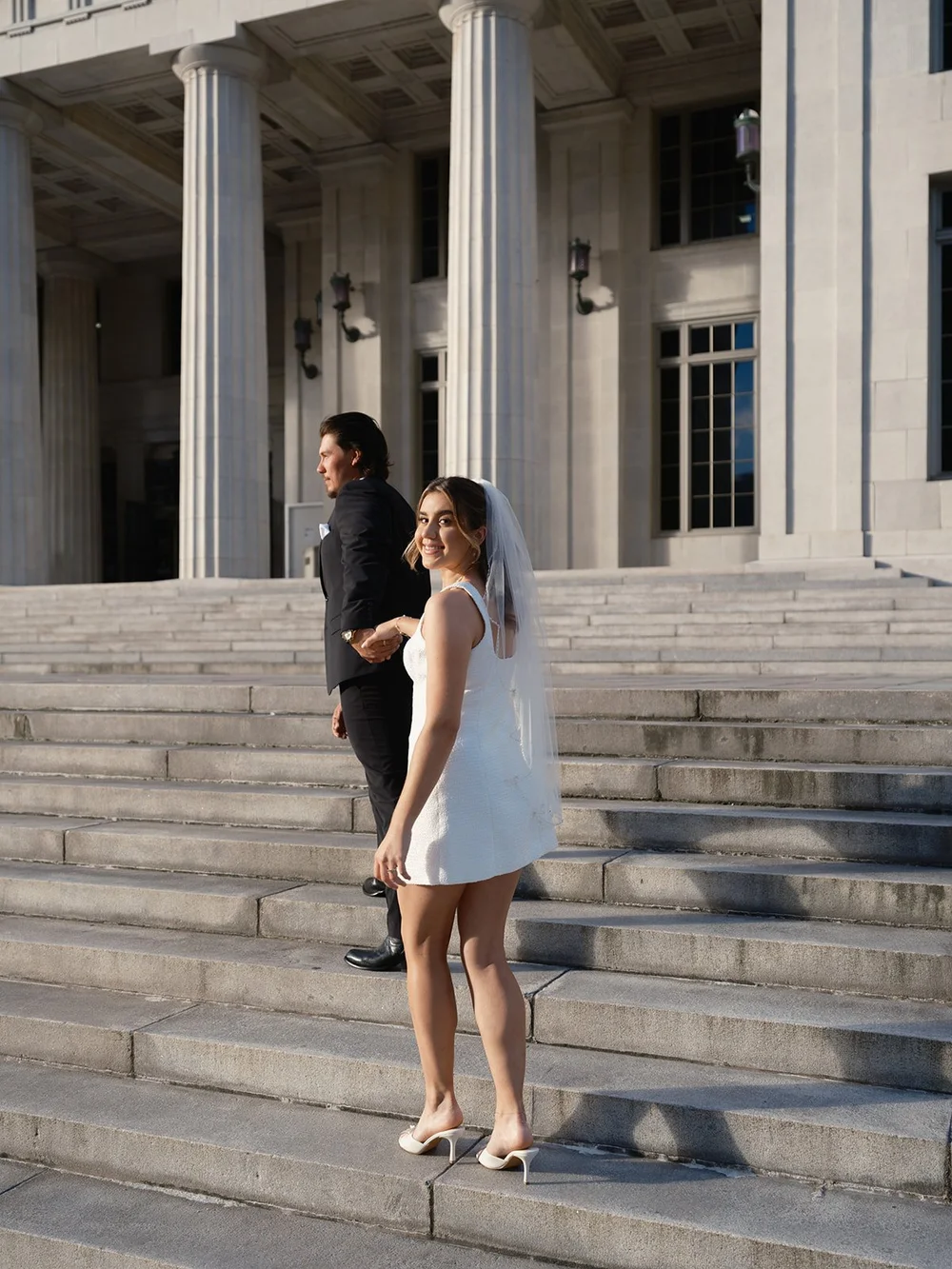 Bride in a short white dress walking down the courthouse steps with the groom behind her