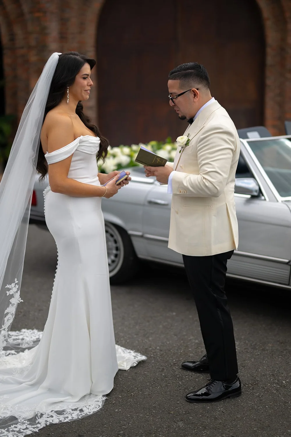 Krystine and Douglas facing each other beside the white vintage car with brick arches behind them