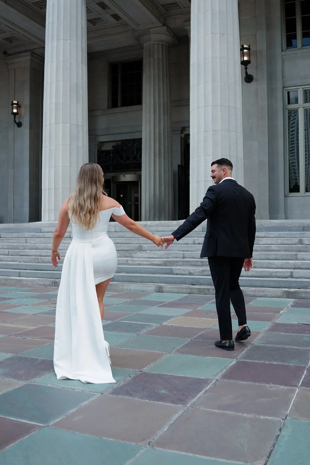 Rachel and Austin walking hand in hand across the colored stone plaza below the courthouse columns