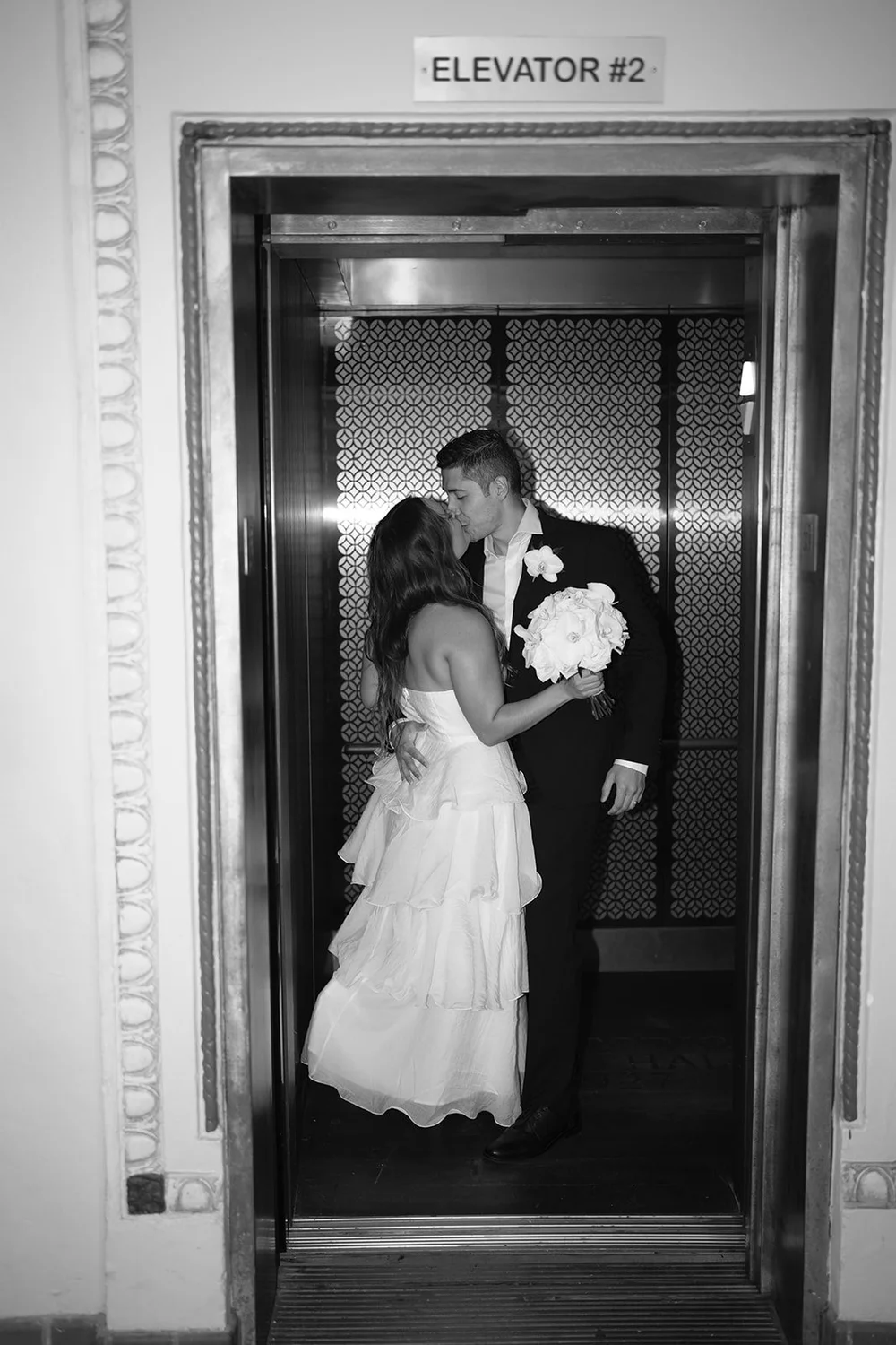 Black and white image of the couple embracing inside an open elevator, the Elevator #2 sign above the door