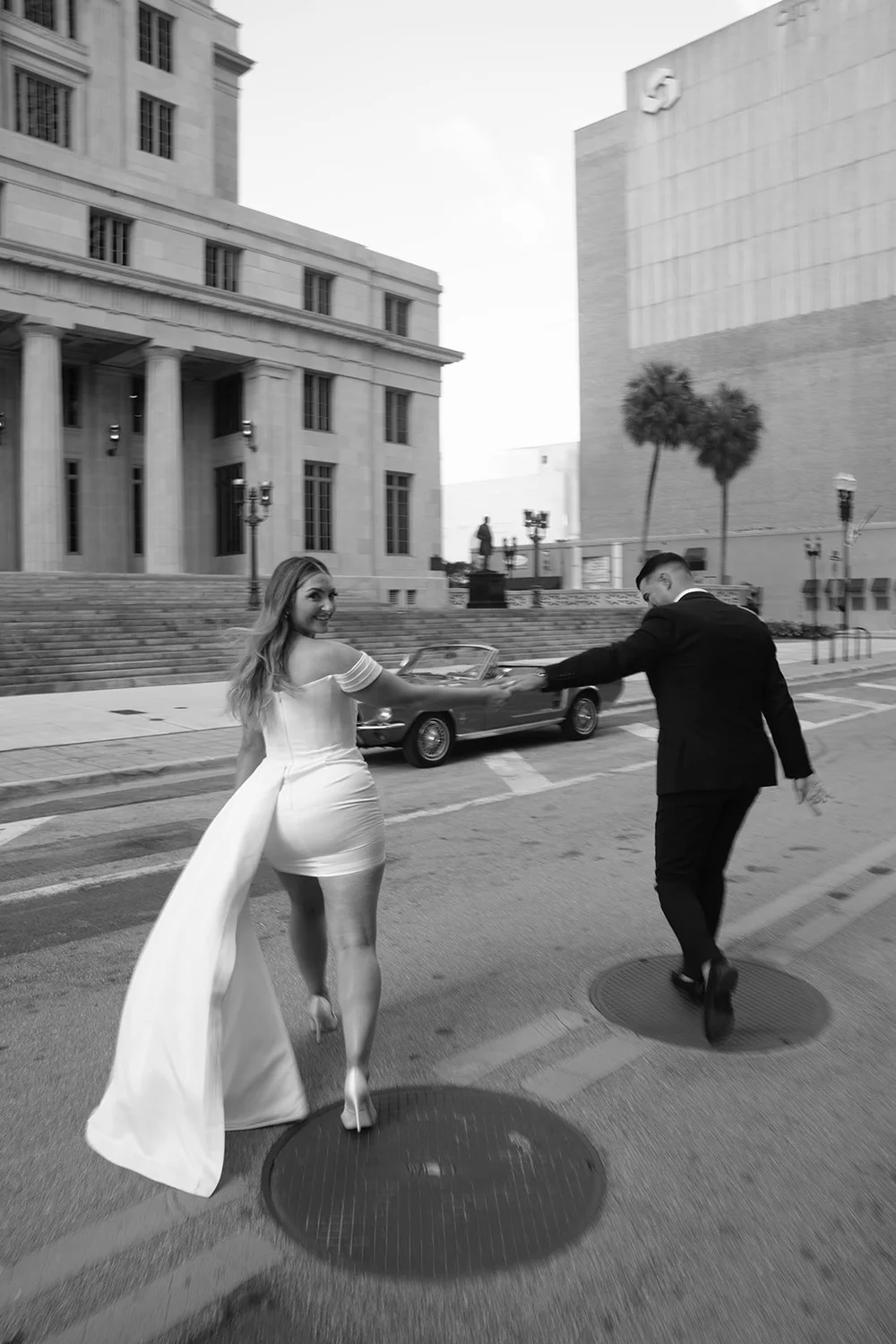 Rachel in a short white dress with a long train walking toward Austin on a downtown street, in black and white