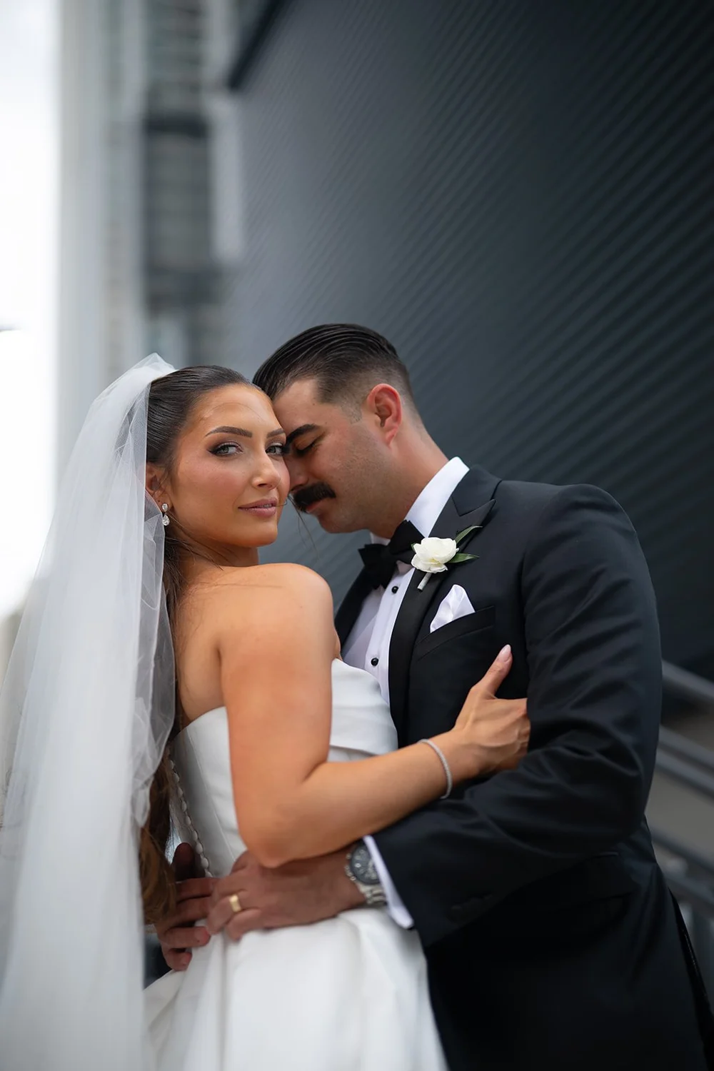Rachel and Austin touching foreheads with her veil around them beside a modern building