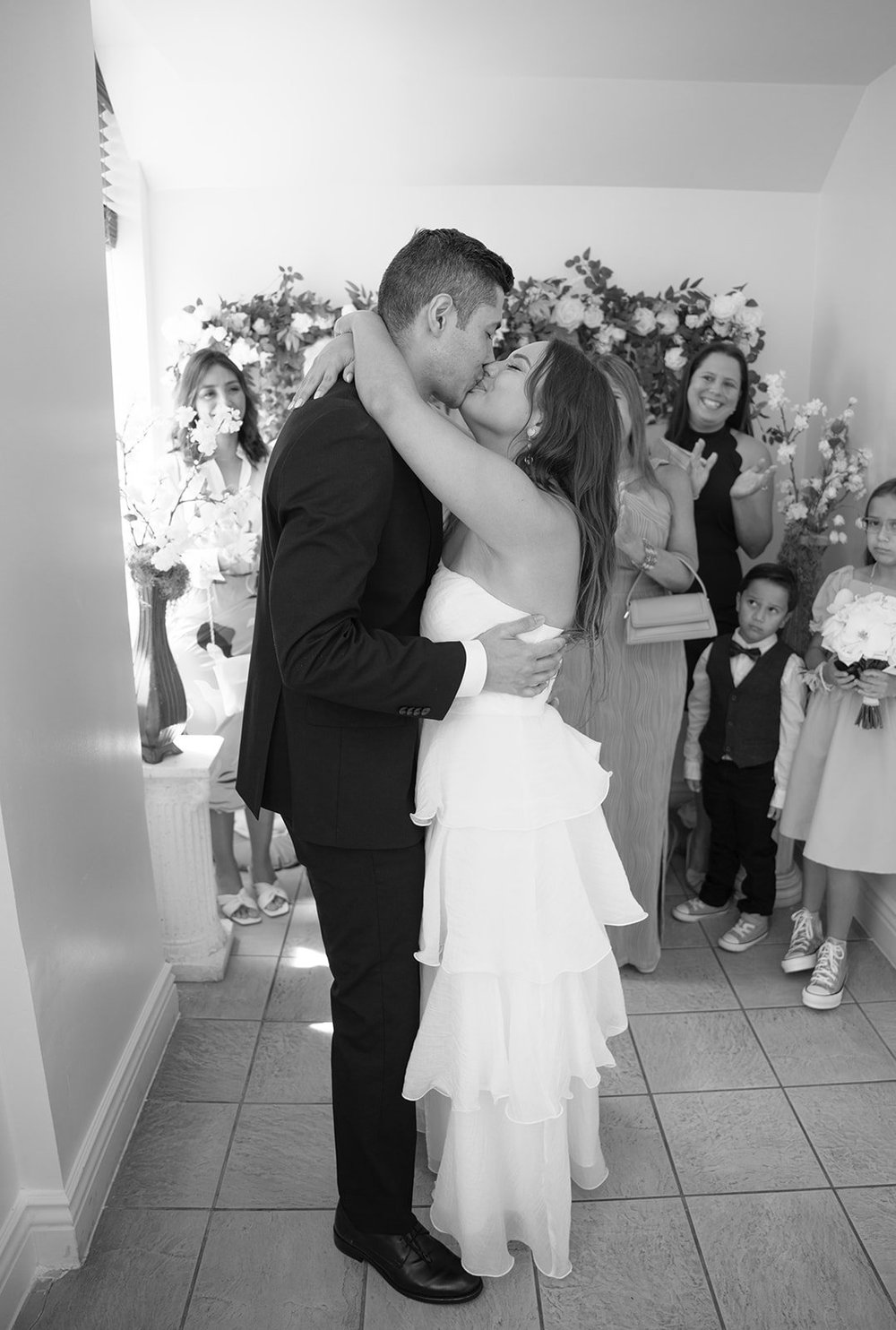 Black and white photo of Chris and David in a close embrace and kiss with the family looking on behind them
