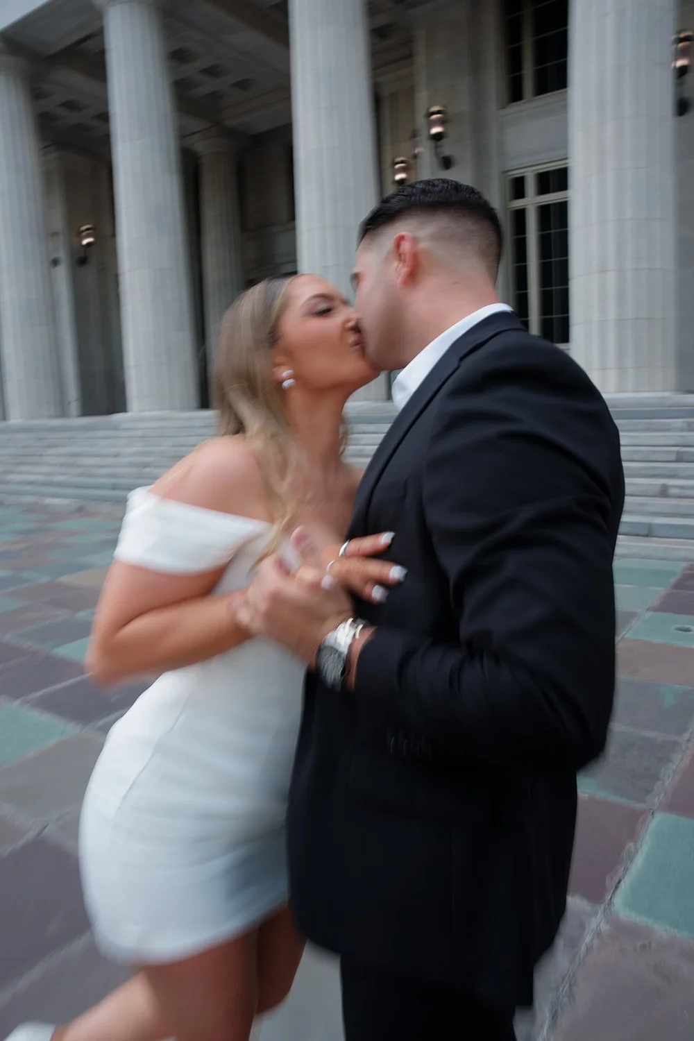 Rachel in a short white dress and Austin in a black suit kissing in a moment of movement near the courthouse columns