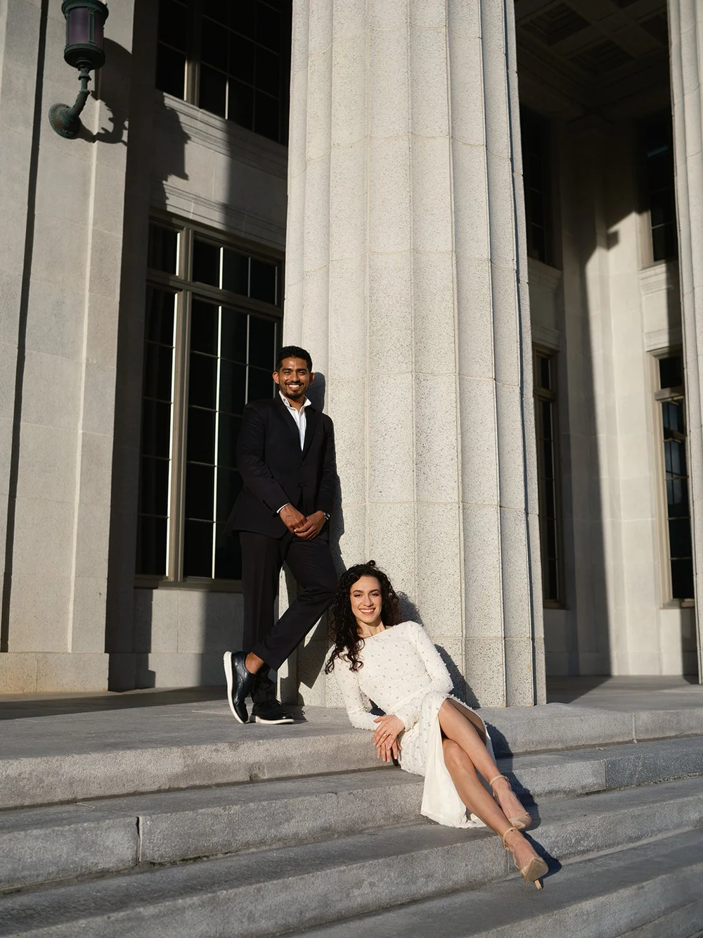 Bride sitting on the courthouse steps while the groom stands beside the tall stone columns