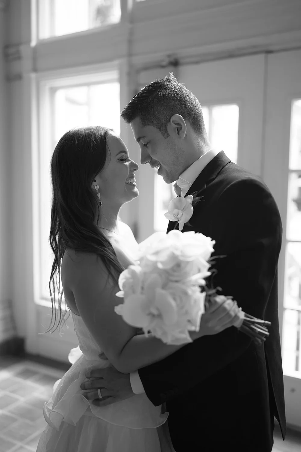 Black and white photo of the couple kissing in front of tall windows, Chris holding her bouquet