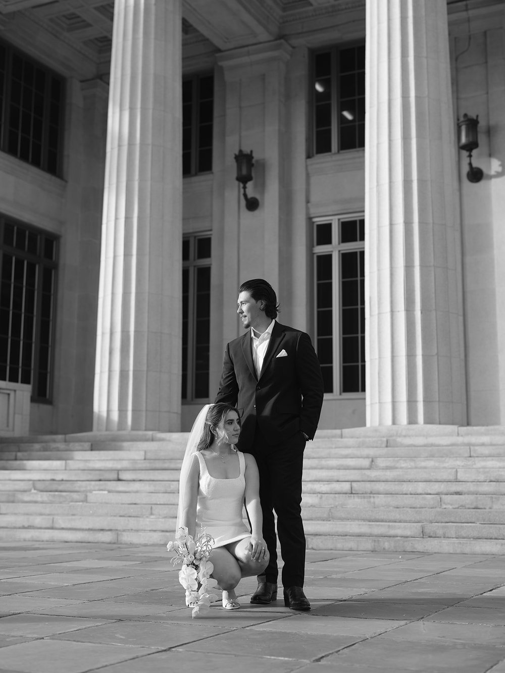 Black and white frame of the bride crouching with her bouquet while the groom stands behind her on the courthouse steps