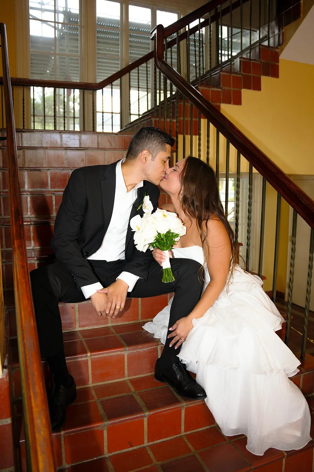 Chris and David kissing as they sit together on a red tile staircase with a dark railing