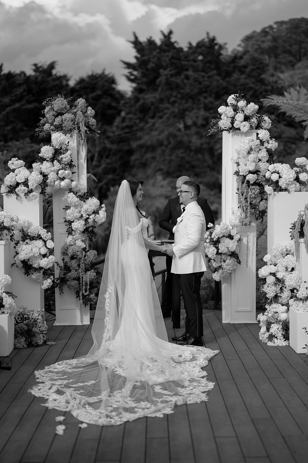 Black and white view of Krystine's long veil trailing down the aisle between white flower arrangements