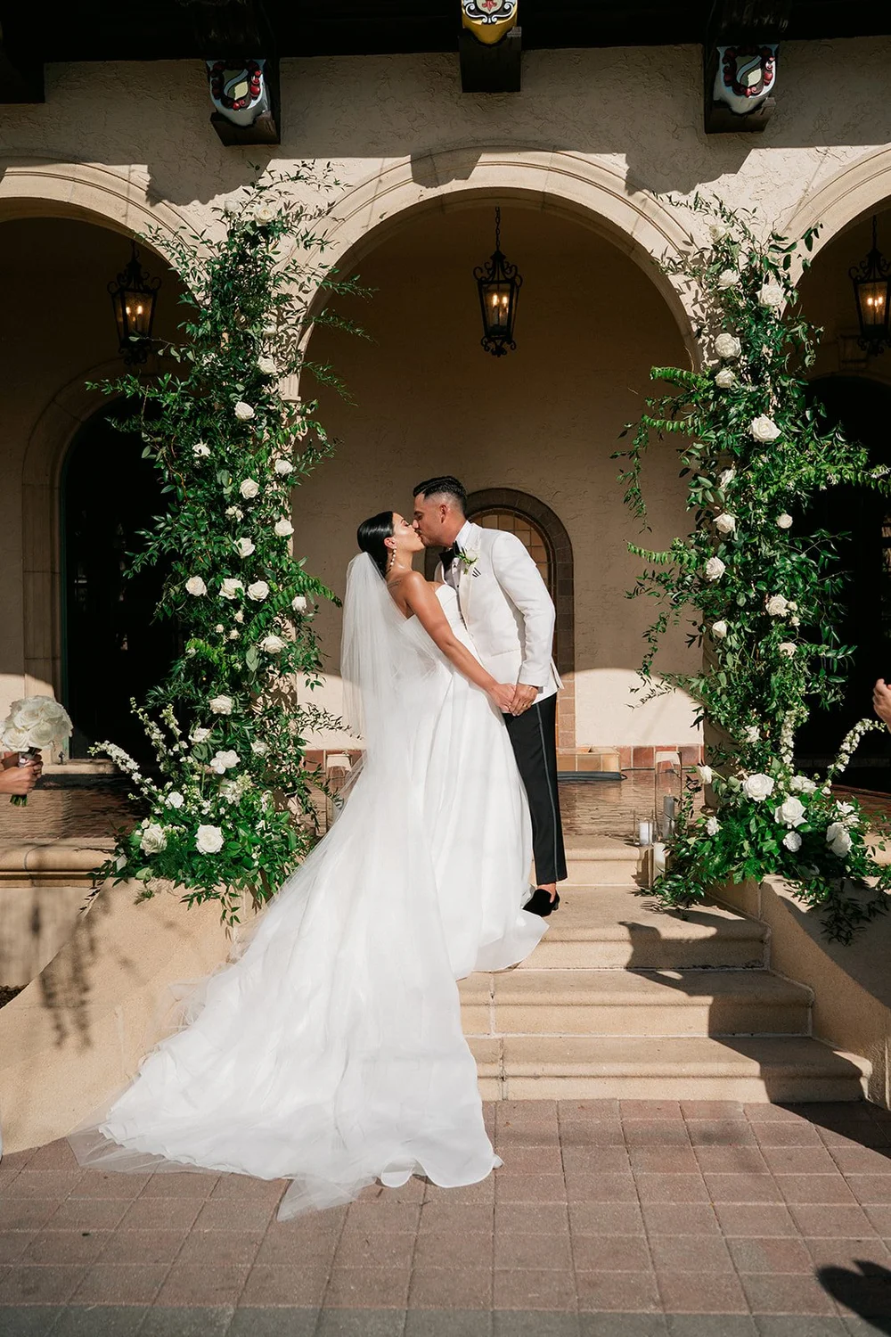 Steph and Ricky kissing under the courtyard arches at the Powel Crosley Estate, framed by columns of white roses and greenery