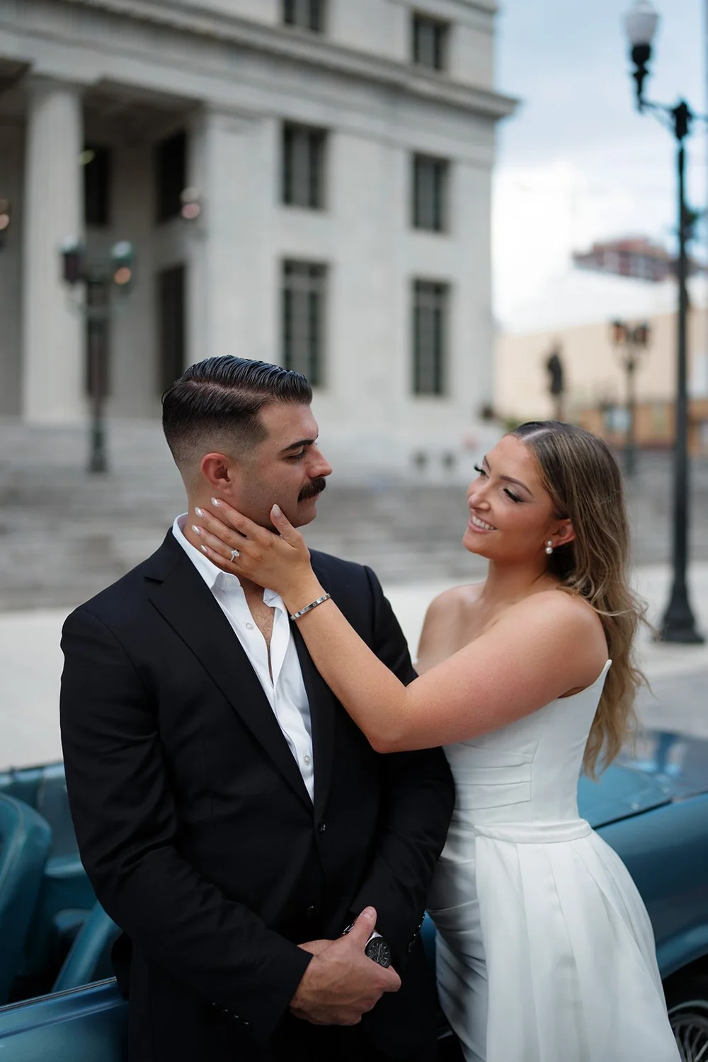 Rachel touching Austin's face as they stand next to the blue Mustang with a street lamp behind them